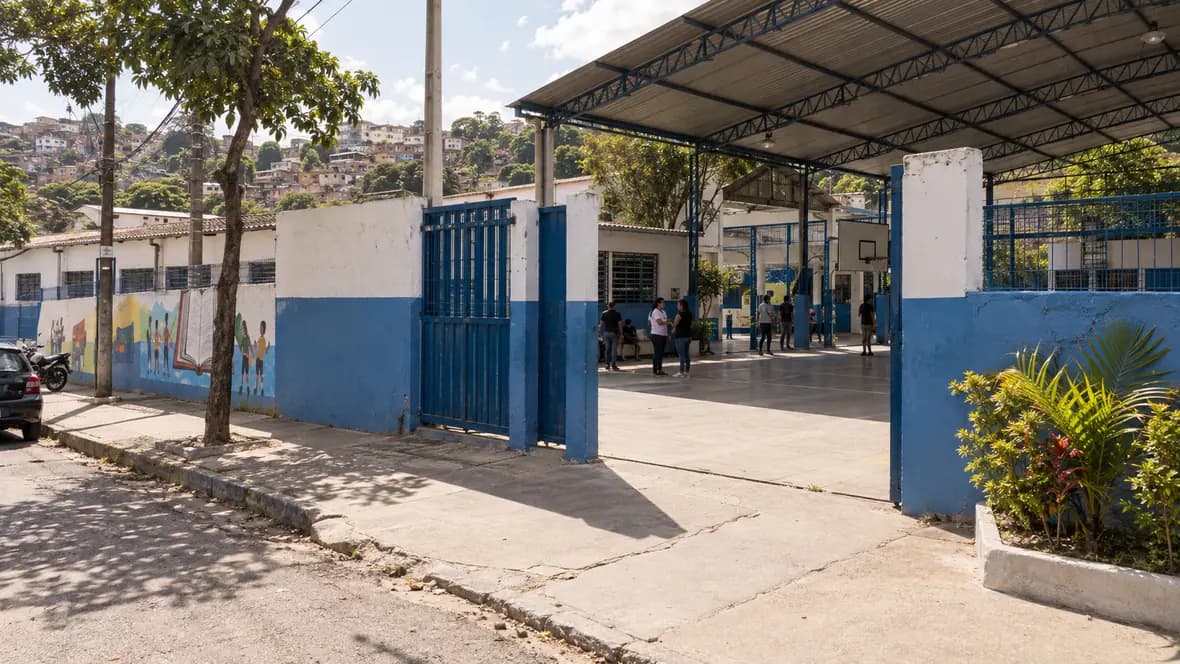 Foto de entrada de escola pública brasileira com adultos circulando em um pátio ensolarado